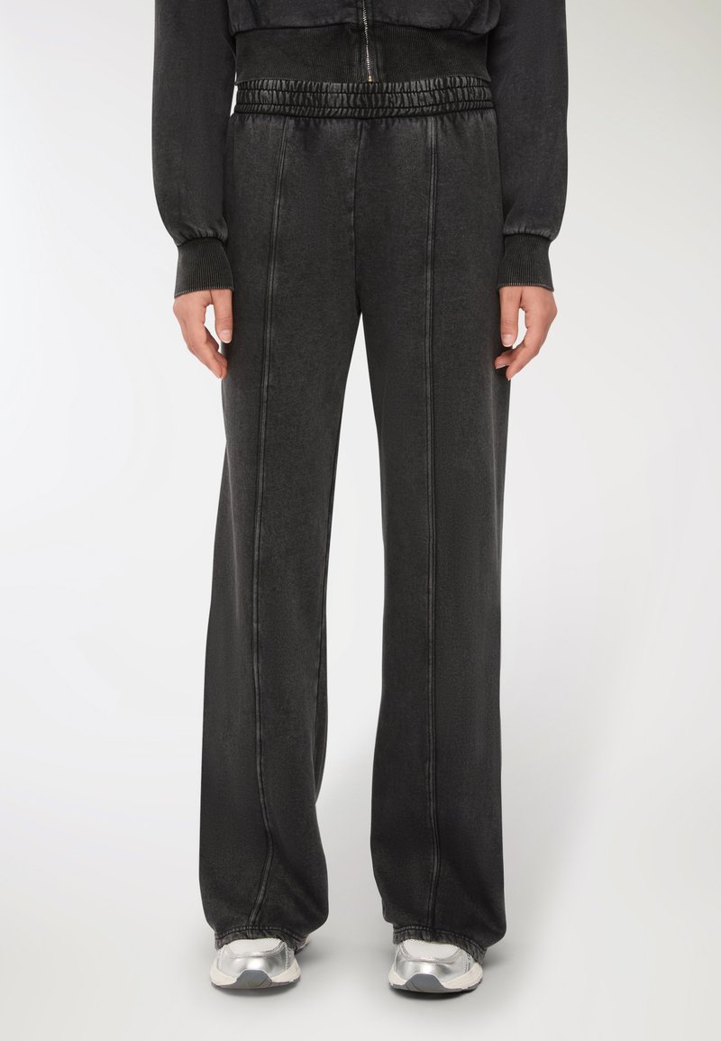 Osoba nosząca luźne czarne dresy z widocznymi detalami szwów oraz elastycznym pasem, w połączeniu z srebrno-białymi trampkami.