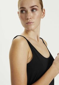 Schwarzes Tanktop mit dünnen Trägern, figurbetontem Design, glatter Textur und tiefem Ausschnitt. Die Schulter und der Hals des Models sind sichtbar, vor einem neutralen Hintergrund.