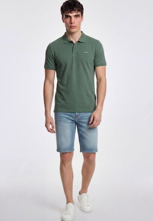 Uomo in piedi indossa una polo verde, pantaloncini di jeans azzurri chiari e sneakers bianche su uno sfondo semplice.