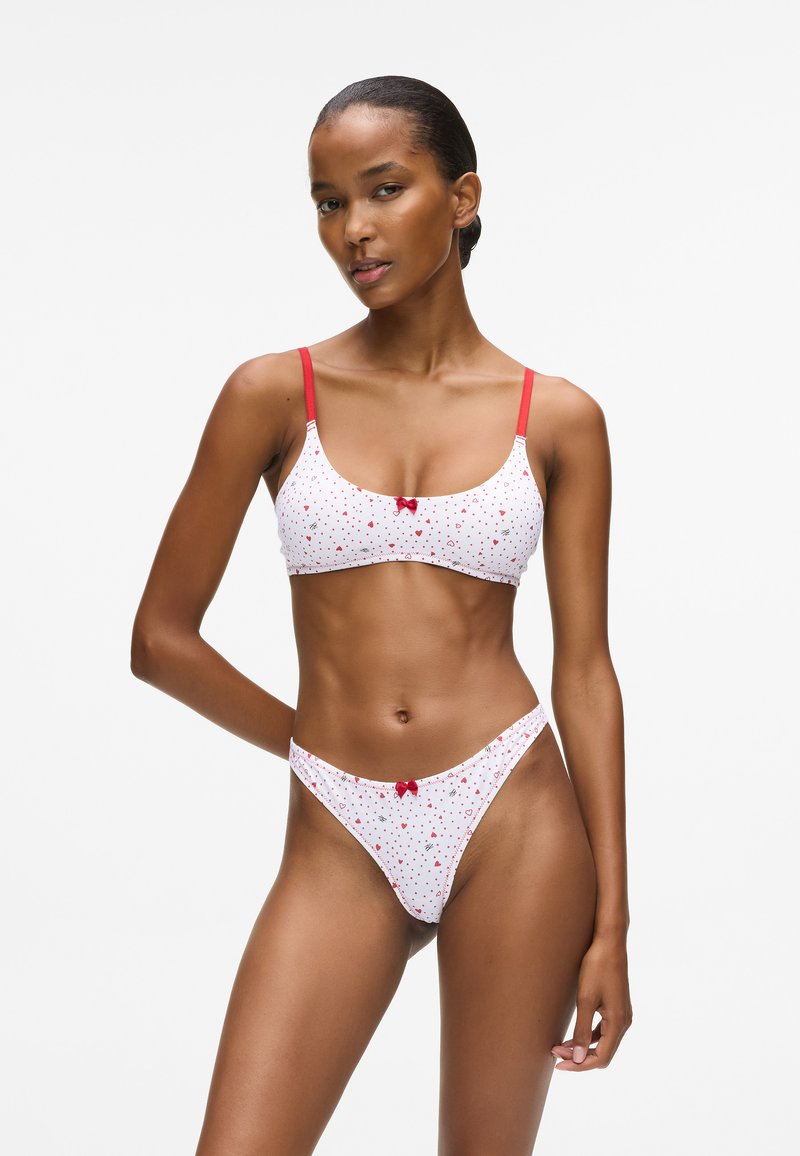 Lieknas moters, vilkinčios baltą ir raudoną širdžių raštuotą liemenėlę bei atitinkamus aukštai iškirptus kelnaites, stojančios prieš paprastą baltą foną.