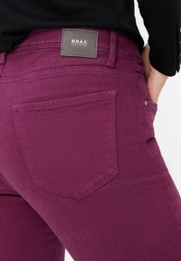 Jeans de mezclilla morados con un diseño ajustado, que cuentan con bolsillos traseros, costuras en contraste y una etiqueta de cuero cuadrada con la inscripción "BRAX".