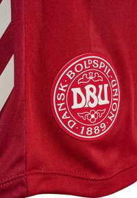 Röd sporttröja med en rund, vit logotyp med "DBU" i centrum, omgiven av blommiga detaljer och texten "DANSK BOLDSPIL-UNION 1889."