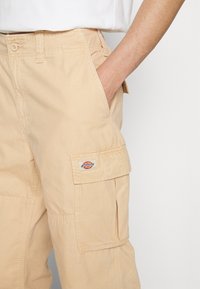 Beige cargo pants made of cotton, featuring a side pocket and a small branded patch. The design includes a button closure and belt loops.