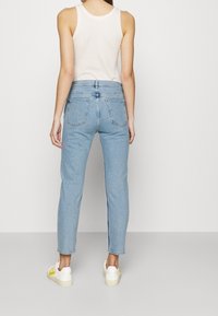 Jean bleu clair taille haute avec une jambe droite, cinq poches et des détails de couture subtils, associé à des chaussures blanches avec des accents jaunes.