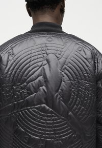 Άτομο που φοράει μαύρο μπουφάν τύπου bomber με κεντητό σχέδιο καλαθοσφαιριστή και περιμετρικό κείμενο στην πλάτη, πάνω σε απλό φόντο.