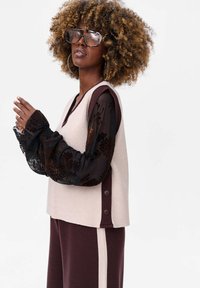 Beżowy top bez rękawów z burgundowymi akcentami, nałożony na czarną przezroczystą koszulę z długim r ękawem w kwiaty; zestawiony z pasującymi burgundowymi spodniami.