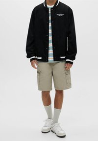 Black suede bomber jacket with white striped cuffs, paired with a multicolored striped shirt, beige cargo shorts, and white sneakers.