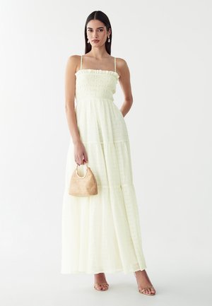 Mujer con un vestido largo amarillo claro con tirantes finos, sosteniendo un pequeño bolso tejido y llevando sandalias doradas y pendientes de perlas.