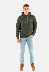 Sudadera verde oliva con cordones, bolsillo frontal y un logo sutil en el pecho, combinada con unos jeans azul claro y botas marrones.