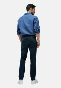 Hombre de pie de espaldas, vistiendo una camisa azul arremangada, jeans oscuros y zapatillas blancas, sobre un fondo gris claro liso.