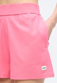 Ροζ σορτς με ελαστική μέση και πλαϊνή τσέπη, φορεμένα με το χέρι μέσα στην τσέπη, με μια μικρή ετικέτα "HUGO" κοντά στο τελείωμα.