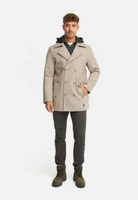 Cappotto doppiopetto beige in misto lana con ampio colletto, cappuccio grigio scuro e due tasche frontali, abbinato a pantaloni scuri e stivali marroni.