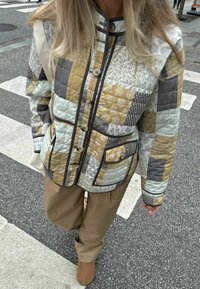 Jachetă matlasată în diverse modele de patchwork în nuanțe de bej, gri și verde mentă; dotată cu nasturi, buzunare și margini contrastante. Pantaloni maro, pantofi bej.