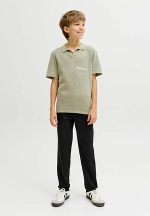 Leende pojke stående med händerna avslappnade, klädd i ljusgrön pikétröja, svarta byxor och vita sneakers mot en enkel ljus bakgrund.