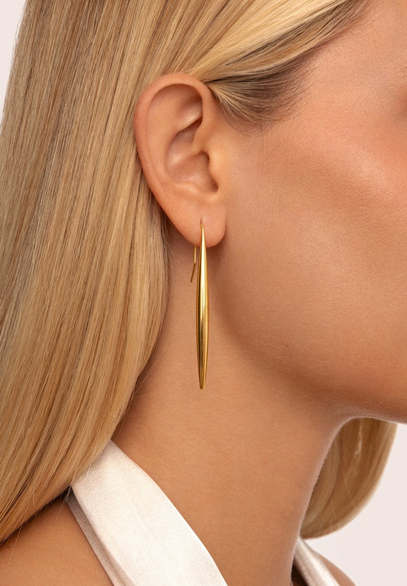 Femme blonde portant une longue boucle d’oreille fine en forme de goutte dorée avec une finition brillante, montrée en gros plan de profil avec une peau lisse et des cheveux raides.