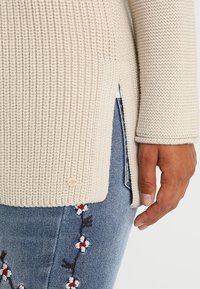 Béžový pletený svetr s žebrovanou texturou, bočními rozparky a dekorativním knoflíkem na lemu. Kombinován s džínovinou s výšivkou.