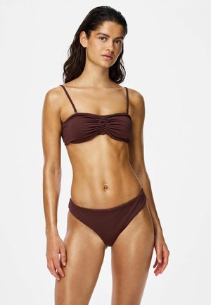 Fiatal nő nedves hajjal, sötétbarna, fodros bandeau felsővel és magasított szabású alsóval rendelkező két darabos bikiniben, egyszínű háttér előtt.