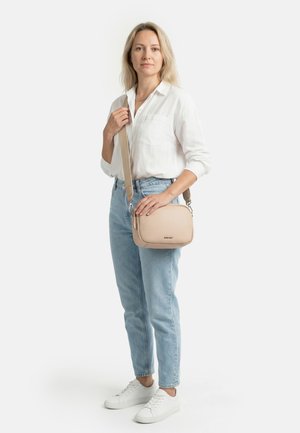 Donna in piedi che indossa una camicia bianca, jeans azzurri chiari, scarpe da ginnastica bianche, che tiene una borsa a tracolla beige con una larga tracolla.