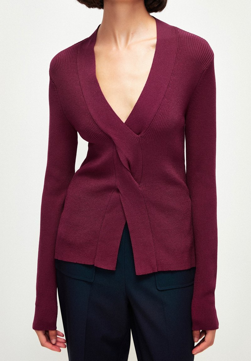 Femme portant un cardigan ajusté côtelé bordeaux avec un col en V profond et des manches longues, associé à un pantalon sombre sur mesure.