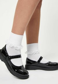 Svarta lackläders Mary Jane-skor med rund tå, rem över foten, i kombination med vita fransiga ankelsockor.