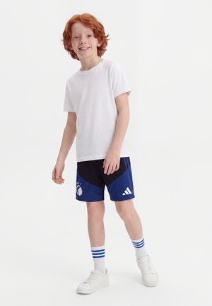 Rødhåret dreng iført en hvid t-shirt, blå og sorte sportsshorts, hvide sokker med blå striber og hvide sneakers, stående mod en ensfarvet baggrund.