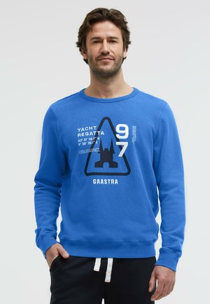 Mann trägt einen blauen Sweatshirt mit dem Schriftzug „Yacht Regatta 97“ und einem schwarzen Emblem, kombiniert mit schwarzen Hosen mit weißen Kordeln.