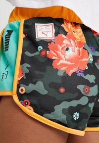 Shorts en camouflage noir avec des motifs floraux vifs en orange et bleu, dotés d'une taille élastique, d'une bordure jaune et d'un patch logo Puma.
