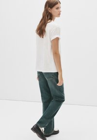 Bela T-maica s podvlečenimi rukavi je v kombinaciji s temno zelenimi ravnimi hlačami. Obutev je črna z belim podplatom, prikazana s hrbtne strani.