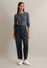 Szary dzianinowy kardigan na guziki, zestawiony z ciemnymi, dopasowanymi spodniami. Modelka ma na sobie białe sneakersy, prezentując elegancki i nowoczesny wygląd.