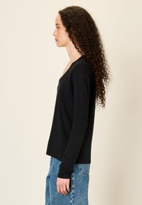 Naine pikkade lokkis juustega, seljas must pikkade varrukatega V-kaelusega top ja sinised teksad, seisab profiilis tavalise tausta ees.