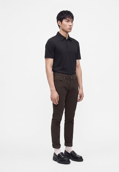 Jeune homme portant un polo noir à manches courtes, un pantalon marron foncé retroussé, des chaussettes blanches et des mocassins noirs, debout devant un fond uni.