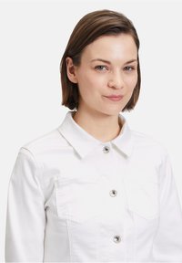 Weißes Jeansjacke mit einem Kragen, zwei Brusttaschen, silbernen Knöpfen und einer strukturierten Passform. Glatte Textur ohne Muster.