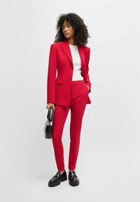 Completo rosso su misura con un blazer strutturato, pantaloni slim-fit e un top bianco a costine. Abbinato a mocassini neri chunky e una piccola borsa.