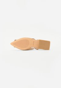 Sandale à talon beige avec un bout pointu, un talon large et une semelle texturée. Elle possède une bride de cheville et un matériau supérieur lisse.