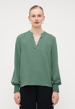 Green, textured blouse with a v-neck and long, gathered sleeves. Cuffs feature smocking detail. Paired with black bottoms.
