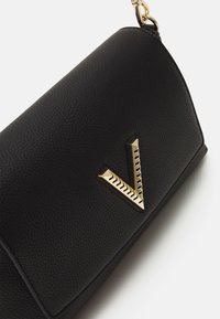 Černá kožená kabelka s texturovaným povrchem, se zlatým kovovým akcentem ve tvaru V. Design je štíhlý a strukturovaný, s řetízkem.