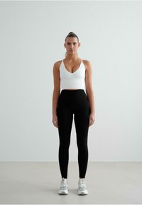 Hvid ribbet cropped top med dyb V-udskæring, parret med højtaljede sorte leggings og grå sneakers. Ensfarvet baggrund.