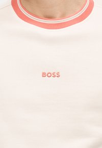 Λευκό μπλουζάκι με στρογγυλή λαιμόκοψη, διακοσμημένο με κοραλλί περίγραμμα και κεντητό logo "BOSS" σε κοραλλί στο κέντρο του στήθους.