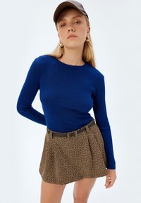 Donna giovane che indossa un maglione a coste blu, una gonna marrone a fantasia con cintura, un berretto grigio e orecchini a cerchio dorati su uno sfondo neutro.
