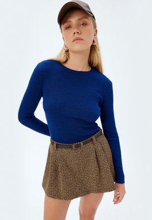 Joven mujer vistiendo un suéter azul acanalado, falda marrón con estampado y cinturón, gorra gris y pendientes de aro dorados frente a un fondo liso.