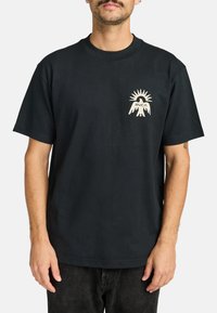 T-shirt in cotone nero con maniche corte, dotato di una grafica crema che rappresenta un sole stilizzato e delle mani sul lato sinistro del petto.