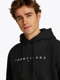 Svart hettegenser laget av bomull, med "TOMMY JEANS" logo i hvit brodering, snorstrenger og en frontlomme som en kenguru.