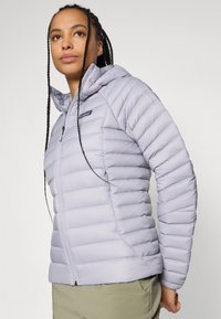 Kvinne med flettet hår iført en lys lilla quilted jakke som ser litt oppover, med hendene bak ryggen, mot en ensfarget bakgrunn.