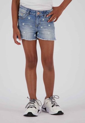 Krótkie spodenki dżinsowe w jasnoniebieskim kolorze z motylkową aplikacją, postrzępionym dołem i klasycznym pięciokieszeniowym krojem. Połączone z białymi trampkami.