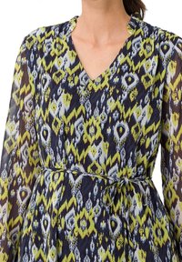 Femme portant une robe à décolleté en V avec de longues manches transparentes et un motif zigzag et losanges multicolore en jaune, marine et blanc, cintrée à la taille.