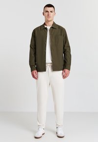 Olivenfarbene Button-up-Hemd mit einer Brusttasche, über einem weißen T-Shirt getragen, kombiniert mit cremefarbenen Jogginghosen und weißen Turnschuhen.
