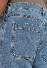 Πίσω όψη ατόμου που φοράει ανοιχτό μπλε τζιν παντελόνι με ορατές πίσω τσέπες και λεπτομέρειες ραφής, σε συνδυασμό με μπεζ μπλούζα.