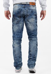 Jean en denim bleu avec un délavage usé, des coutures décoratives, deux poches arrière et une étiquette en cuir brun sur la taille. Jambes légèrement fuselées.