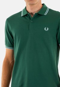 Polo verde hecho de tela texturada con una tapeta de dos botones, acentos de doble raya blanca en el cuello y un pequeño logotipo bordado.