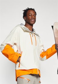 Ski jacket in cream with orange and grey accents. Features zip pockets, adjustable hood, and elastic cuffs. Smooth, weather-resistant fabric.